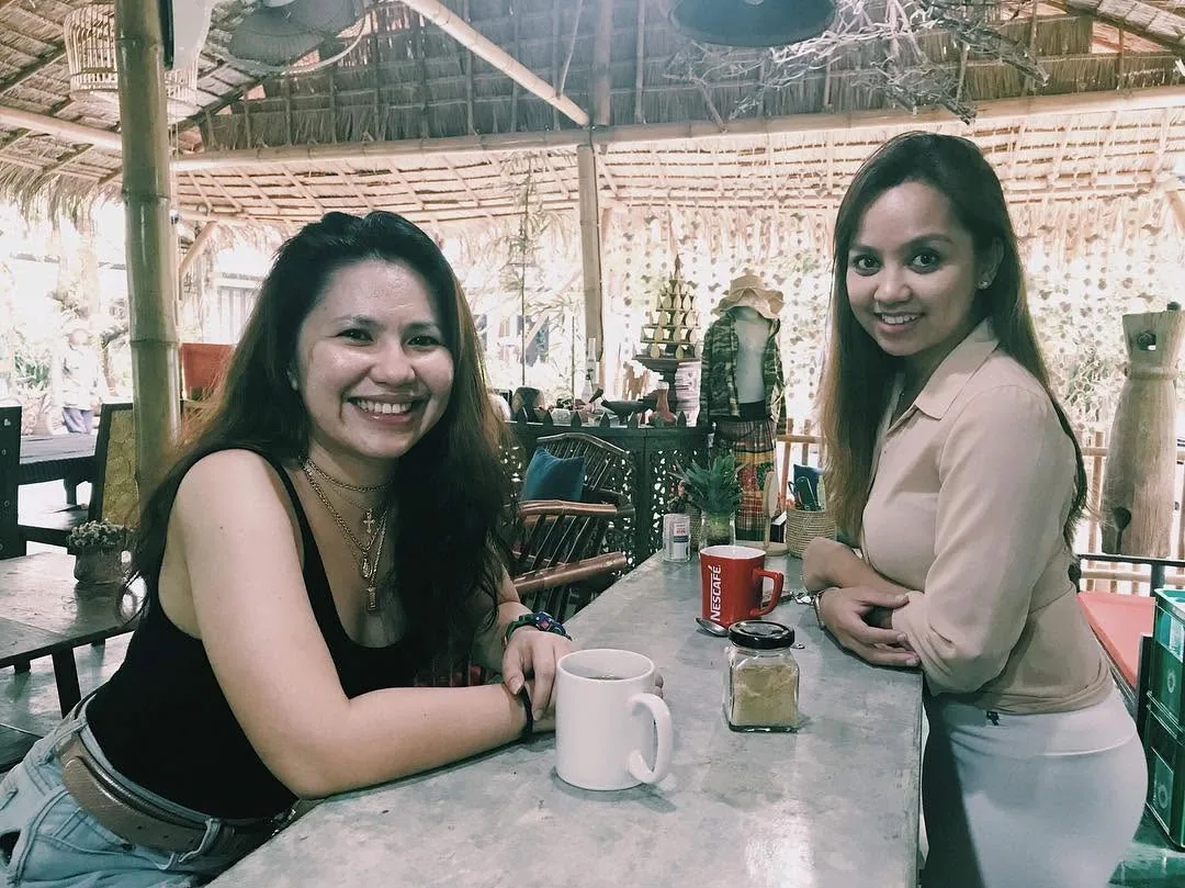 Dee and Kimy smiling over coffee at a thatched cafe table