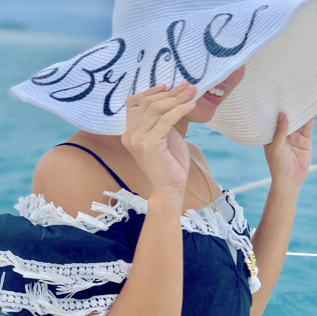 Dee smiling under a wide white sun hat with Bride written across the brim, turquoise sea behind
