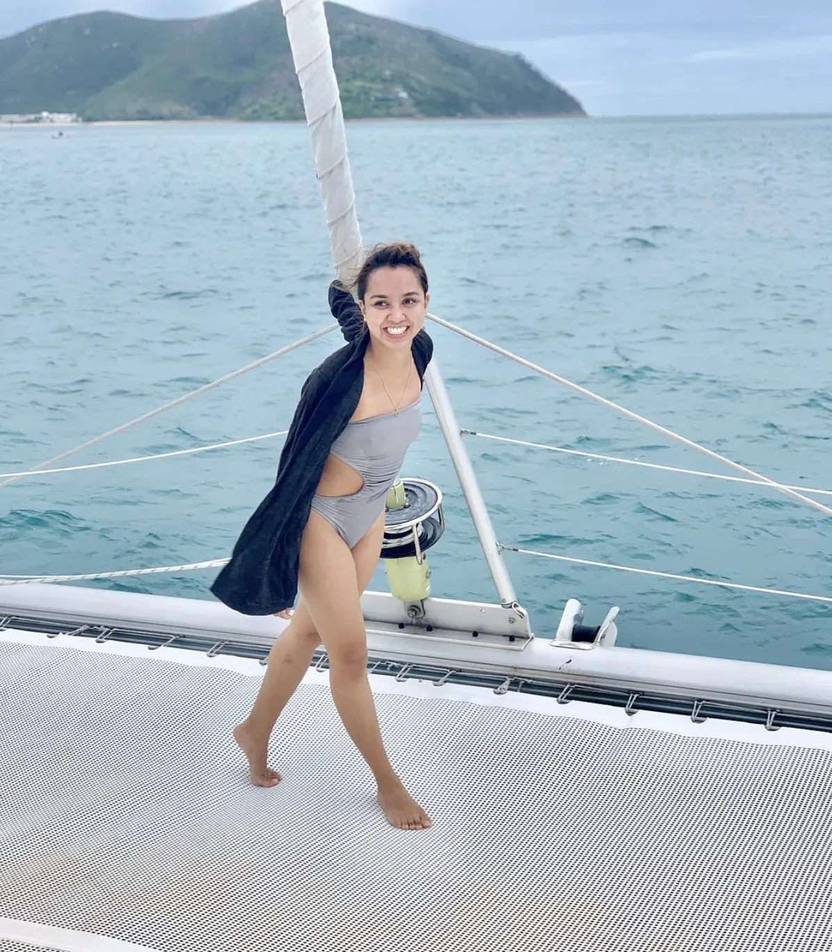 Dee smiling as she walks barefoot across the net of a catamaran, island and sea behind