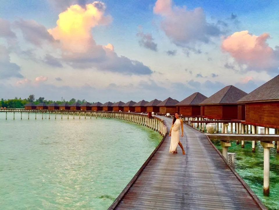 Dee in a white dress on a wooden boardwalk lined with overwater villas at sunset