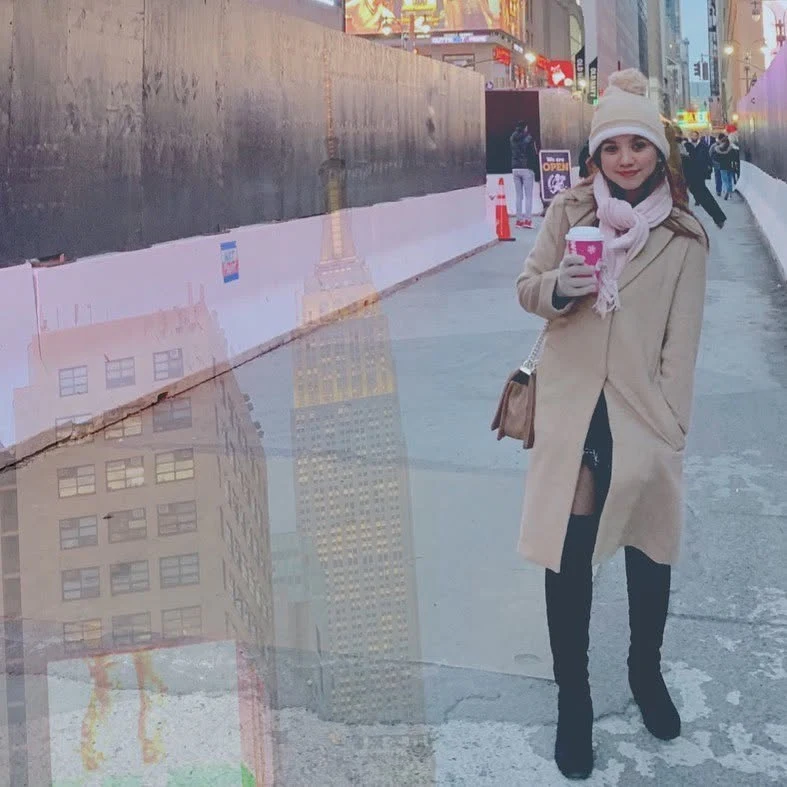 Dee in a camel coat, cream bobble hat and pink scarf, holding a red holiday cup on a Manhattan street, with the Empire State Building layered over the scene