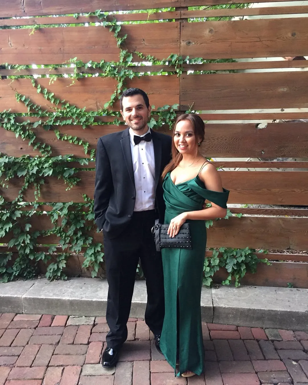 Dee in a green gown and her husband in a tuxedo by an ivy-covered fence