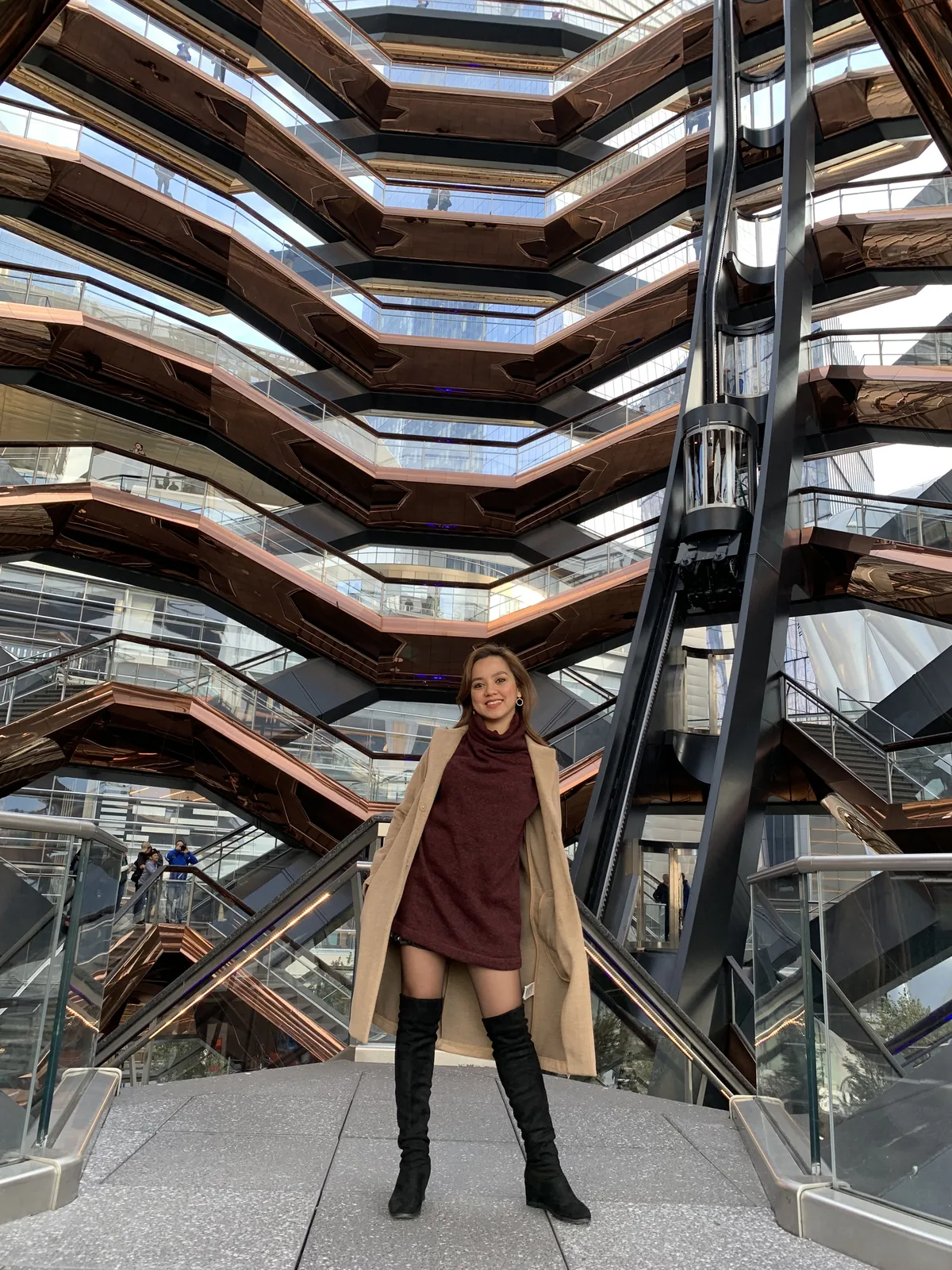 Dee in a camel coat and over-the-knee boots beneath the copper staircases of the Vessel