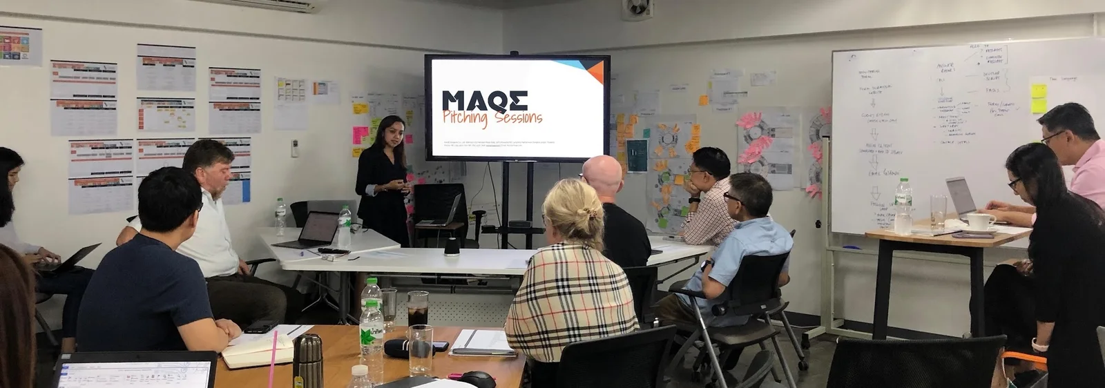 Dee presenting at a MAQE pitching session, with colleagues around the table and walls covered in sticky notes, journey maps and a whiteboard flow